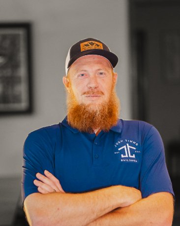 Man in a blue Josh Timms Builders shirt and cap, arms crossed, representing custom homebuilding vision.